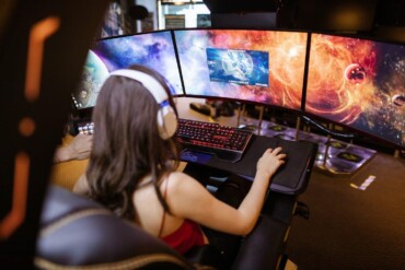 Free Woman with headphones playing on a multi-screen gaming setup featuring vibrant space imagery. Stock Photo