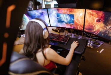 Free Woman with headphones playing on a multi-screen gaming setup featuring vibrant space imagery. Stock Photo