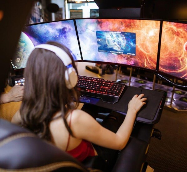 Free Woman with headphones playing on a multi-screen gaming setup featuring vibrant space imagery. Stock Photo